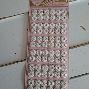 May include: A vintage card of off-white, circular, thread-covered buttons arranged in rows. The pink paper backing features the brand name Thuringia, the text 1a. Kranzknöpfe, and the measurement 20'''. The buttons have a radial, woven pattern.