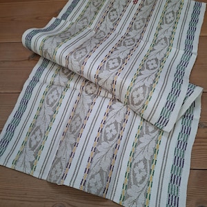 Puede incluir: Un textil plegado con un patrón blanco y gris con rayas verticales y motivos florales. El textil tiene detalles en verde, amarillo y morado. La tela está doblada sobre una superficie de madera.