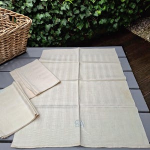 May include: Cream-coloured linen napkins and a large tablecloth with a subtle striped pattern. The tablecloth has the embroidered initials "SA" in the corner. A wicker basket is in the background, suggesting a rustic or vintage theme.