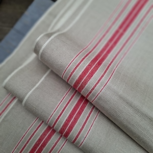 Könnte beinhalten: Ein gefaltetes Stück hellbeiger Stoff mit einer Reihe vertikaler Streifen in leuchtendem Rot und dünnen weißen Linien. Das Textil weist eine sichtbare Webstruktur auf und ist mehrlagig, um das sich wiederholende Muster zu zeigen.