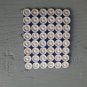 May include: A rectangular blue card holding forty-eight circular buttons arranged in eight rows of six. Each button features a light beige, thread-wrapped surface with a radiating pattern of fine lines converging at the center of the button.