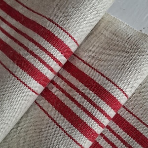 Könnte beinhalten: Nahaufnahme von gefaltetem Leinenstoff mit natürlichem beigem Hintergrund und kräftigen roten Streifen. Der Stoff hat ein strukturiertes Gewebe, und die Streifen verlaufen diagonal über das Material. Die Falten erzeugen Schatten und verleihen dem Bild Tiefe. Dieser Stoff eignet sich für verschiedene Dekorationsprojekte.