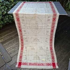 Könnte beinhalten: Cremefarbene Leinentischdecke mit rotem Rand und Akzenten. Die Tischdecke hat ein zentrales Design und sich wiederholende Muster. Das Wort "Rolltuch" ist unten rot aufgedruckt. Die Tischdecke ist über einen grauen Tisch drapiert.