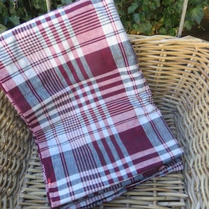 Op de afbeelding: Een rood-wit geblokt tafelkleed met een bordeauxrood en grijs ruitpatroon. Het tafelkleed is gevouwen en in een rieten mand gelegd.