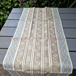 May include: A rectangular table runner featuring a woven pattern of beige floral vines and leaves on a cream background. The design includes vertical stripes in blue and white, with a small red embroidered detail near the top center edge.