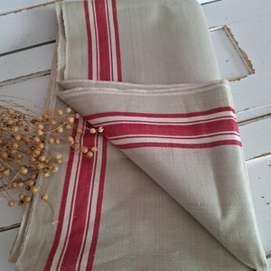 Könnte beinhalten: Gefaltetes Küchenhandtuch aus Leinen in Naturbeige mit kräftigen rot-weißen Streifen. Die Streifen verlaufen vertikal und horizontal über den Stoff. Das Handtuch hat eine rustikale, ländliche Ästhetik.