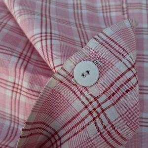 May include: A light pink and white plaid fabric garment featuring a single round white button centred on a folded cuff. The textile displays a repeating grid pattern of thin red and white lines intersecting across a soft pink background.
