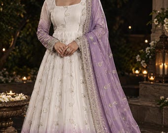 May include: A floor-length white gown featuring long sleeves, a square neckline, and a lavender ombre hemline. The dress is adorned with silver floral embroidery and sequins. A matching lavender dupatta with silver borders drapes over the shoulder.