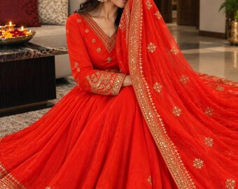 May include: A vibrant red floor-length Anarkali dress featuring intricate gold embroidery and mirror work along the hem, sleeves, and neckline. The garment includes a matching sheer red dupatta with gold floral motifs and a wide embellished border.