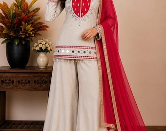 May include: A beige three-piece outfit consisting of a long-sleeved tunic with a red heart-shaped chest panel, wide-leg palazzo pants, and a sheer red dupatta. The tunic and pants feature silver mirror work, red embroidery, and small white beads.