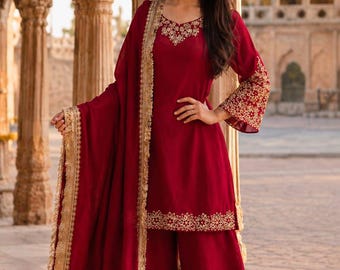 May include: A deep red three-piece outfit consisting of a long-sleeved tunic, wide-leg trousers, and a matching dupatta. The tunic features gold floral embroidery around the neckline, sleeve cuffs, and hem, while the dupatta has a gold fringe border.