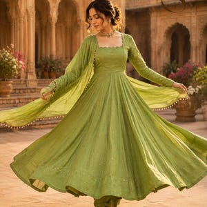 May include: A light green Anarkali dress with long sleeves, a square neckline, and a flared skirt. The outfit includes a matching sheer dupatta with small decorative beads along the edges. The fabric features a subtle, shimmering texture.