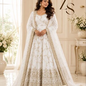 May include: A floor-length white Anarkali dress featuring intricate gold embroidery and sequin detailing throughout the bodice and skirt, paired with a matching sheer dupatta with a wide gold embroidered border. The letters ZS appear in the top right.