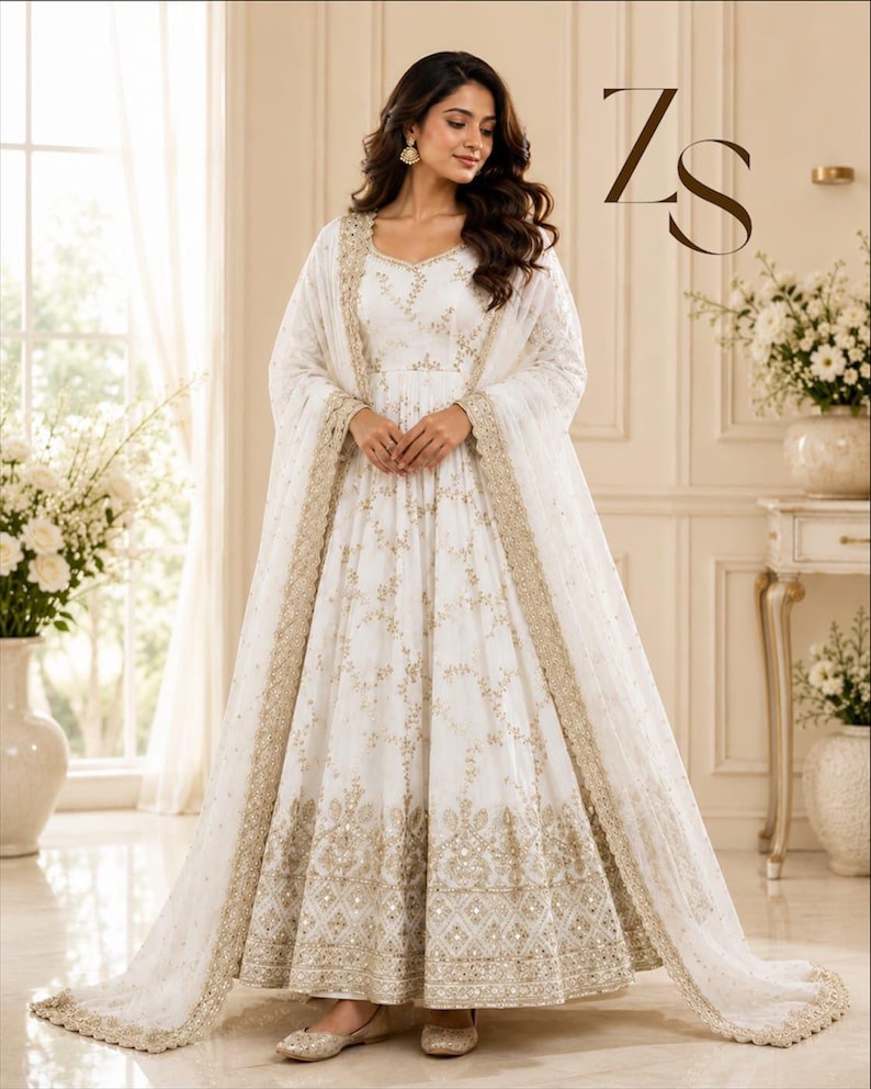 May include: A floor-length white Anarkali dress featuring intricate gold embroidery and sequin detailing throughout the bodice and skirt, paired with a matching sheer dupatta with a wide gold embroidered border. The letters ZS appear in the top right.