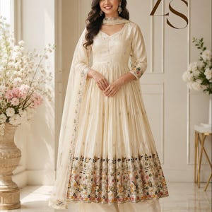 May include: A cream-colored Anarkali suit featuring a V-neckline, three-quarter sleeves, and a flared skirt with a multi-colored floral embroidered border. The ensemble includes a matching dupatta and wide-leg trousers, with the letters Zs visible.