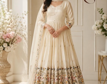 May include: A cream-colored Anarkali suit featuring a V-neckline, three-quarter sleeves, and a flared skirt with a multi-colored floral embroidered border. The ensemble includes a matching dupatta and wide-leg trousers, with the letters Zs visible.