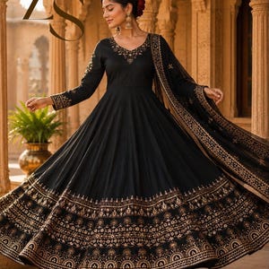May include: A black floor-length Anarkali dress featuring long sleeves, a V-neckline, and intricate gold embroidery along the bodice, cuffs, and hem. The flared skirt displays detailed metallic patterns, accompanied by a matching sheer dupatta.