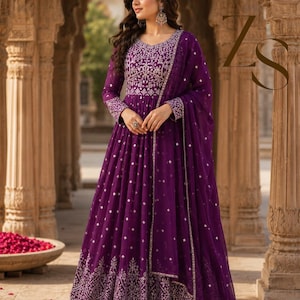 May include: A floor-length deep purple Anarkali dress featuring intricate silver embroidery on the bodice and hemline. The dress includes long sleeves and a matching sheer purple dupatta with small silver accents, creating a cohesive look.