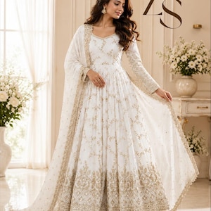 May include: A floor-length white gown featuring intricate gold embroidery and sequin detailing throughout the bodice and sleeves. The skirt is pleated with a heavy gold embroidered border at the hem, paired with a matching sheer white dupatta.