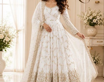 May include: A floor-length white gown featuring intricate gold embroidery and sequin detailing throughout the bodice and sleeves. The skirt is pleated with a heavy gold embroidered border at the hem, paired with a matching sheer white dupatta.