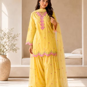 May include: A bright yellow three-piece outfit consisting of a long-sleeved tunic, loose-fitting trousers, and a sheer dupatta. The tunic features a V-neckline with pink heart-shaped appliqués, scalloped pink hem edges, and small pink heart embroidery.