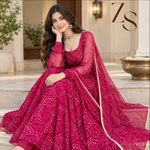 May include: A vibrant magenta floor-length dress featuring a white tie-dye pattern, long sheer sleeves with gathered cuffs, and a scoop neckline. The garment includes a matching magenta dupatta with a gold-toned border and the letters ZS.