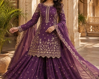 May include: A deep purple sharara suit featuring a long-sleeved tunic with a round neckline and intricate gold embroidery. The matching wide-leg pants and sheer dupatta are adorned with gold floral patterns and scalloped edges throughout.