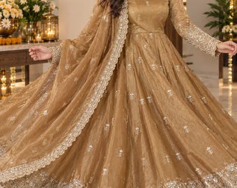 May include: A gold-colored floor-length gown with long sleeves, featuring intricate silver embroidery and sequin detailing throughout the skirt and bodice. The dress includes a matching sheer dupatta with a scalloped, embellished border.
