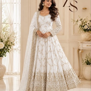 May include: A white floor-length Anarkali dress featuring intricate gold embroidery and mirror work throughout the bodice, sleeves, and flared skirt. The garment includes a matching sheer dupatta with a heavy gold embroidered border and ZS logo.