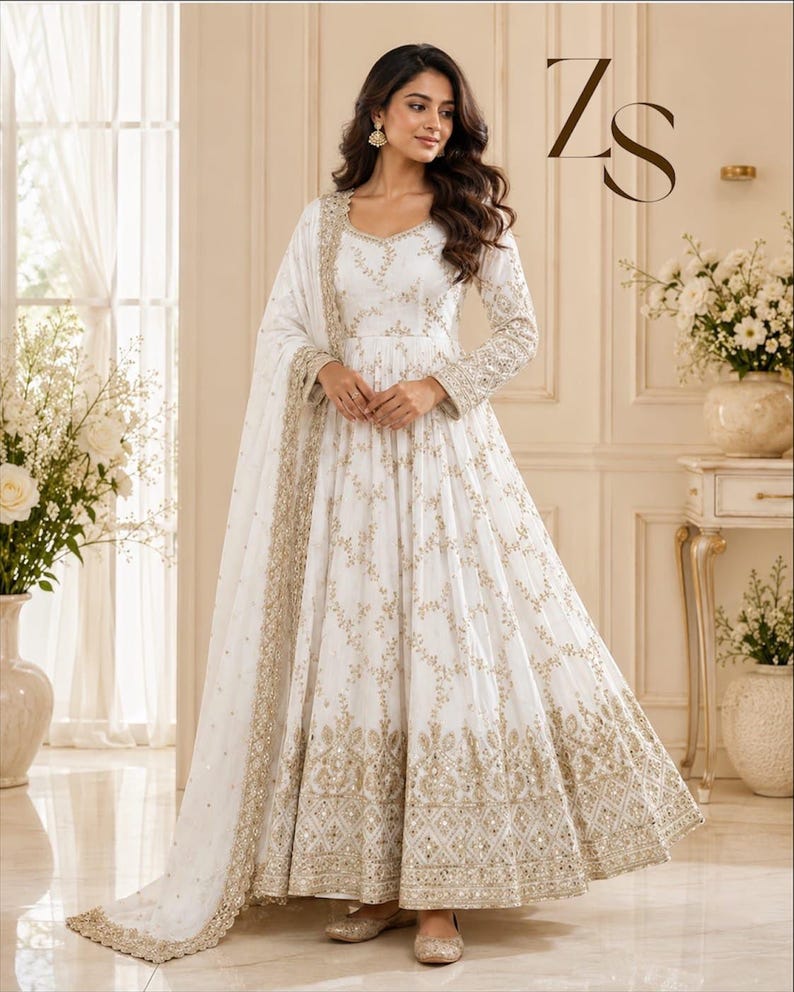 May include: A white floor-length Anarkali dress featuring intricate gold embroidery and mirror work throughout the bodice, sleeves, and flared skirt. The garment includes a matching sheer dupatta with a heavy gold embroidered border and ZS logo.