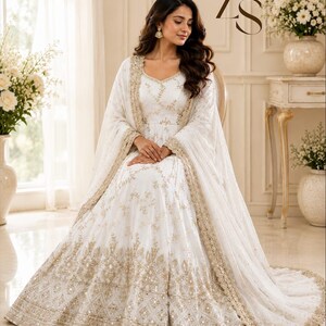 May include: A white floor-length gown featuring intricate gold embroidery and sequin detailing throughout the bodice and skirt, paired with a matching sheer dupatta with a scalloped gold border. The ZS logo is visible in the upper right corner.