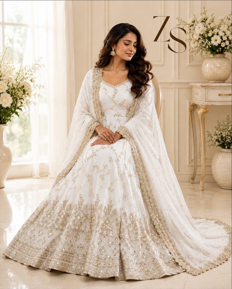 May include: A white floor-length gown featuring intricate gold embroidery and sequin detailing throughout the bodice and skirt, paired with a matching sheer dupatta with a scalloped gold border. The ZS logo is visible in the upper right corner.