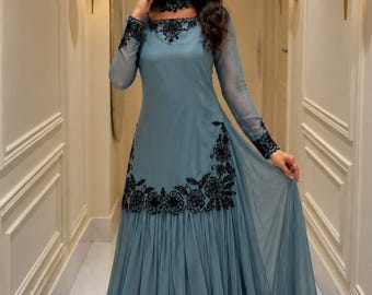 May include: A floor-length slate blue gown featuring long sheer sleeves, a high neckline, and black floral embroidery detailing around the collar, cuffs, and waistline. The skirt is pleated and flows to the floor, crafted from a lightweight fabric.