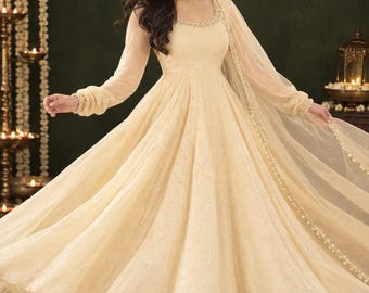 May include: A floor-length cream-colored Anarkali dress with long sleeves, a scoop neckline, and intricate embroidery. The dress features a flared skirt and a matching sheer dupatta with a beaded border. The letters ZS are visible in the top right corner.