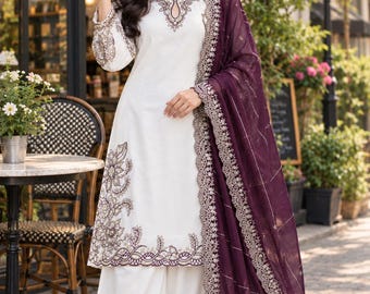 May include: A white long-sleeved tunic and wide-leg trousers set featuring dark purple and gold floral embroidery along the neckline, sleeves, and hem, paired with a matching dark purple sheer dupatta with an embroidered border.