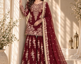May include: A deep maroon sharara suit featuring intricate gold embroidery on the long-sleeved tunic and wide-legged trousers. The ensemble includes a matching sheer maroon dupatta with a gold embroidered border and small gold floral motifs.