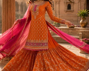 May include: An orange long-sleeved tunic with intricate gold embroidery and a pink border, paired with a matching flared skirt featuring a gold floral pattern. A sheer pink dupatta with gold trim drapes across the outfit, which features the letters LS.