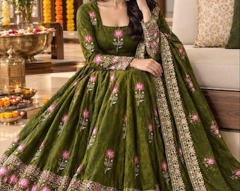 May include: An olive green floor-length dress with a square neckline and long sleeves, featuring an embroidered pink lotus flower pattern throughout. The hem and sleeve cuffs are adorned with a wide, intricate border of pink lotus motifs.