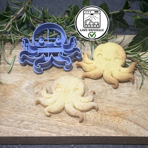 Peut inclure: Un emporte-pièce en plastique violet en forme de pieuvre accompagné de deux biscuits en forme de pieuvre. Une icône blanche circulaire avec un symbole de lave-vaisselle noir et le texte LAVE-VAISSELLE est visible au-dessus.