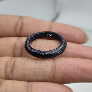 Può includere: Un anello circolare liscio realizzato in pietra arenaria blu scuro con una finitura scintillante e maculata che ricorda un cielo stellato. L'interno della fascia presenta le lettere CGA chiaramente stampate in un piccolo carattere bianco.