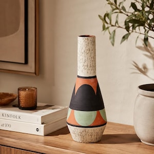 May include: A tall, slender vase featuring a textured cream-coloured neck and base with a speckled finish. The central body displays a geometric pattern of black arches and sage green semi-circles set against a burnt orange background.