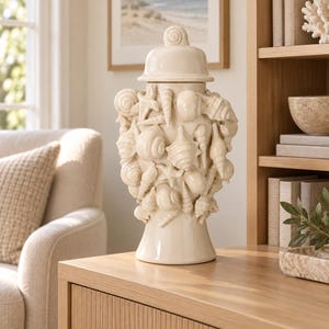 Peut inclure: Un pot en céramique crème avec couvercle, doté d'un extérieur texturé orné de divers coquillages et étoiles de mer sculptés. Le récipient présente une finition lisse et brillante ainsi qu'un couvercle assorti avec un petit bouton en spirale.
