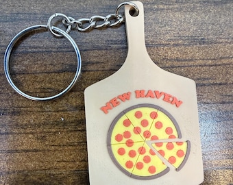 May include: A beige keychain shaped like a pizza peel featuring a yellow pizza with red pepperoni slices and the text NEW HAVEN in orange letters. The keychain includes a silver metal split ring attached by a short silver metal chain.