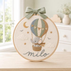 Puede incluir: Un bastidor de bordado circular con un dise&ntilde;o cosido de un elefante gris en un globo aerost&aacute;tico con un lazo verde en la parte superior. La escena incluye una luna creciente, estrellas, nubes y p&aacute;jaros, con el nombre "milo" bordado en cursiva.