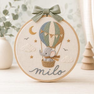 Puede incluir: Un bastidor de bordado circular con un globo aerost&aacute;tico cosido y un elefante gris en la cesta. El dise&ntilde;o incluye una luna creciente, estrellas, nubes y el nombre "milo" en letra cursiva. Un lazo de tela verde salvia est&aacute; sujeto arriba.