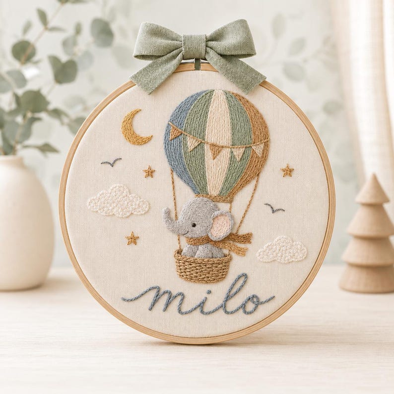 Puede incluir: Un bastidor de bordado circular con un globo aerost&aacute;tico cosido y un elefante gris en la cesta. El dise&ntilde;o incluye una luna creciente, estrellas, nubes y el nombre "milo" en letra cursiva. Un lazo de tela verde salvia est&aacute; sujeto arriba.