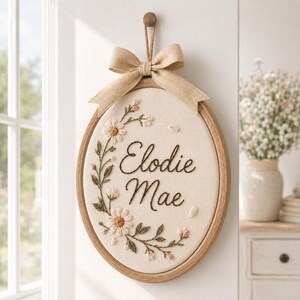 May include: An oval wooden embroidery hoop containing cream-colored fabric with the embroidered name Elodie Mae in dark brown script. The design features a floral vine with white daisies and green leaves, topped with a tan burlap bow.