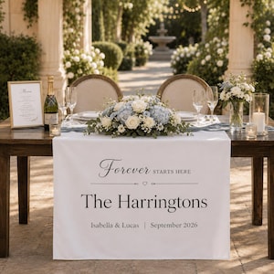May include: A white wedding table runner featuring the printed text Forever STARTS HERE, The Harringtons, Isabella & Lucas, and September 2026. The runner is draped over a wooden table set with a floral centerpiece, wine glasses, and a menu.