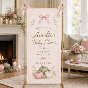 May include: A vertical rectangular banner on a gold metal stand with pink bows at the top corners. The banner features the text "Welcome to Amelia's Baby Shower, so happy you're here! July 24, 2026" with illustrations of pink bows, leaves, and three pumpkins.