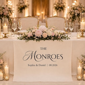May include: A white wedding table runner featuring the black printed text THE MONROES, Sophia & Daniel, and 08.2026. The runner is decorated with a central floral arrangement of pink and white flowers, glass candle holders, and wine glasses.